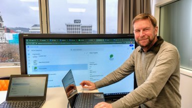 Grant standing in front of the DCH Directory screen with his laptop on the desk next to him, turned to the camera smiling.