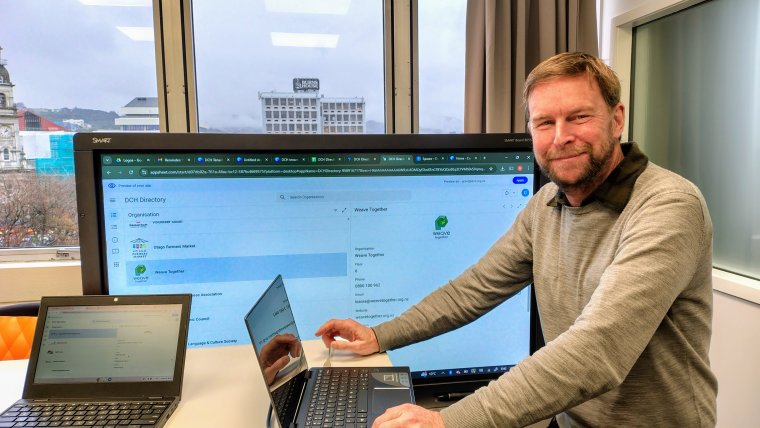 Grant standing in front of the DCH Directory screen with his laptop on the desk next to him, turned to the camera smiling.