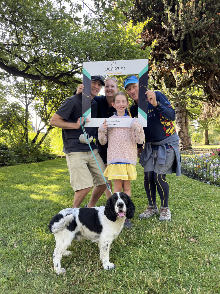 4 people (2 male, 1 female, 1 child) hold up a parkrun frame, black and white dog in foreground