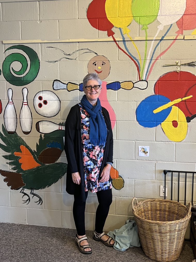 A photo of Sally standing in front of a cinder block wall with paintings of birds, bowling pins, and kiwi toys