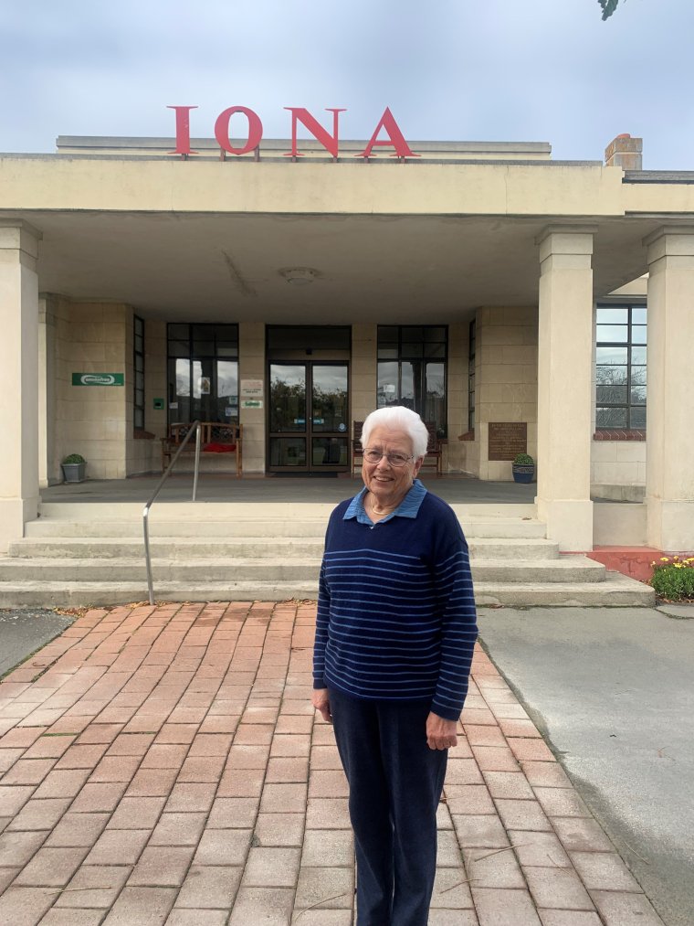 Volunteer Pat standing and smiling in front of the entrance to Iona Care Home