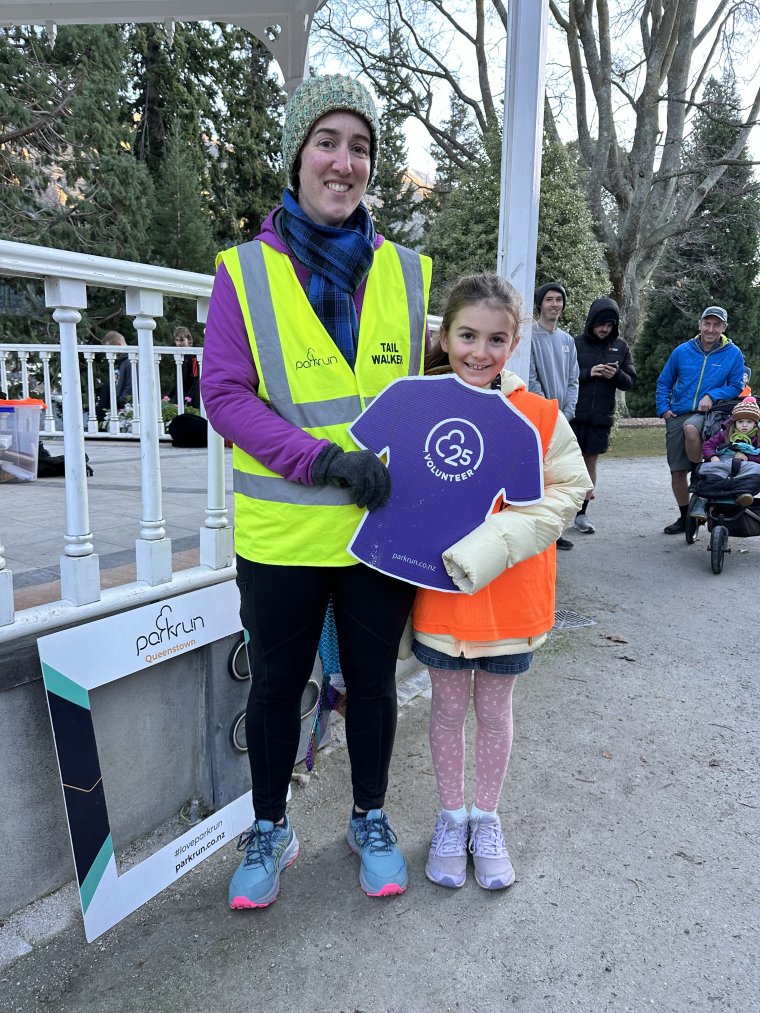 Lisa standing with her daughter, each holding onto one side of a cardboard cutout of a Parkrun volunteer T-shirt and smiling at the camera.