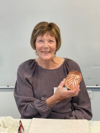 Jan holding up her artistic creation that looks like a browned leaf and smiling at the camera.