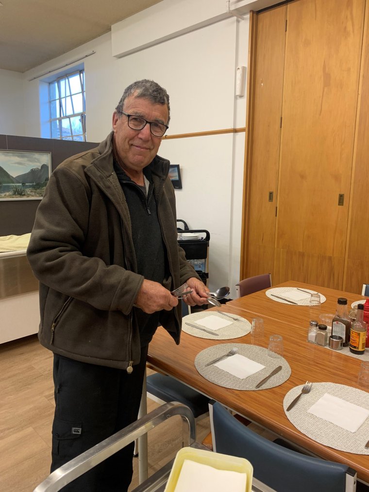 Colin Squire standing next to a set dining table, holding some cutlery, smiling at the camera.