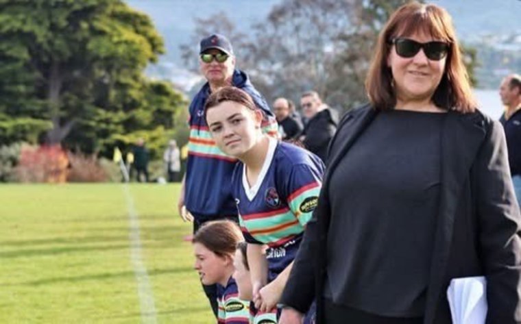 Paula stood on the sidelines of a rugby game, with a few players watching the game beside her.