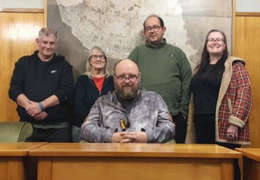 Group of four volunteers stood behind one volunteer sitting at a desk in front of them, all smiling at the camera.