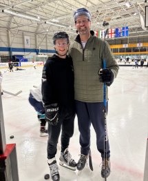 Mat and Toby in ice hockey gear on the ice, smiling at the camera.