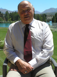 Bruce sitting in a char infront of a lake with mountains in the background. Wearing a striped shirt and red tie.