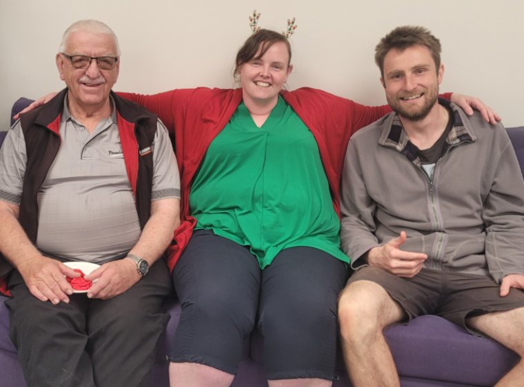 Three volunteers sat together on a couch, the woman in the middle has her arms around those on either side of her and she's wearing a Christmas headband.