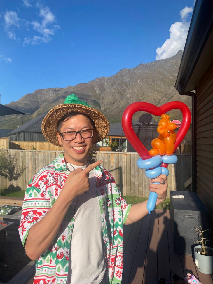 Eric Lim smiling and holding up a balloon animal with a balloon heart around it, pointing at it with his free hand.