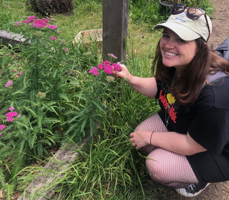 Leigh crouched down smiling at the camera, holding on to one of the flower heads growing in the garden next to her.