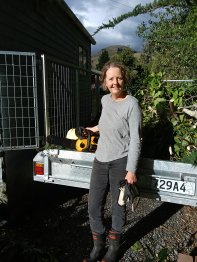 Annabel smiling at the camera, standing outside in her gumboots in front of a trailer full of green waste holding a chainsaw.