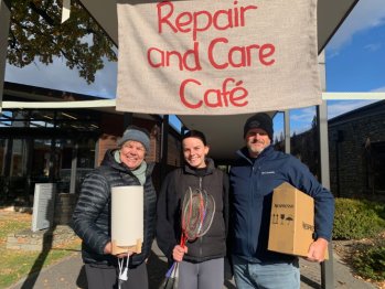 Three people standing outside under the Repair and Care Cafe banner, each holding items to be fixed (a lamp, badminton rackets, a Nespresso machine in a box), smiling at the camera.
