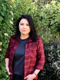 A photo of Monika wearing a red checkered shirt over a blue tshirt, standing in front of a tree.