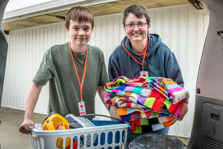 Jasmin holding a knitted blanket and another Red Cross volunteer holding a laundry basket with household supplies, both smiling at the camera.