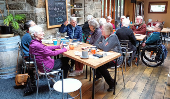 A group of people sitting at two tables, having coffee in a cafe, engaged in conversation.